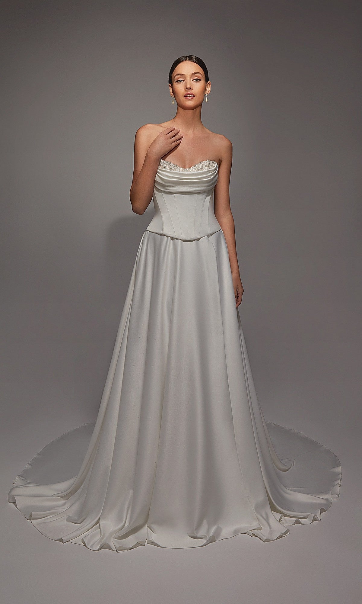 A woman wears the Alyce 7142, a long white dress with a corset bodice and flowing train, its satin fabric softly shimmering against a plain gray background.